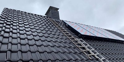 Ein Dach mit glänzenden schwarzen Ziegeln, auf dem eine Leiter und ein Solarpanel montiert sind, unter einem bewölkten Himmel.