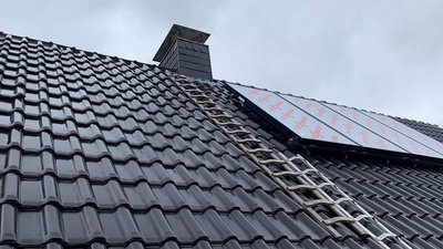 Ein Dach mit glänzenden schwarzen Ziegeln, auf dem eine Leiter und ein Solarpanel montiert sind, unter einem bewölkten Himmel.