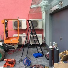 Eine orangefarbene Hebebühne steht neben einer Leiter und mehreren Klimaanlagen an einer pinken Wand.