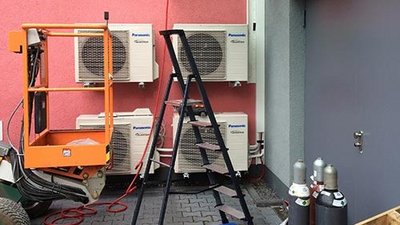 Eine orangefarbene Hebebühne steht neben einer Leiter und mehreren Klimaanlagen an einer pinken Wand.