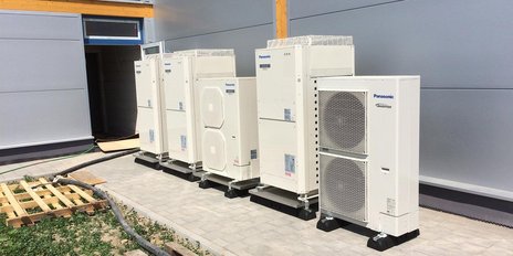 Fünf große Klimaanlagen stehen nebeneinander auf einem gepflasterten Boden vor einer modernen Gebäudeaußenwand.