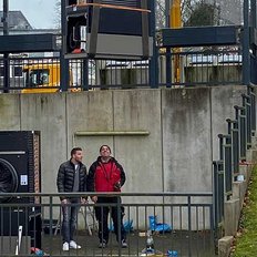 Zwei Männer stehen an einer Baustelle und beobachten einen großen Kran, der eine schwere Last hebt.