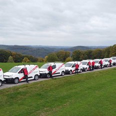 Eine Reihe von Lieferwagen steht auf einer Wiese, während Mitarbeiter in roten Jacken auf dem Weg posieren.