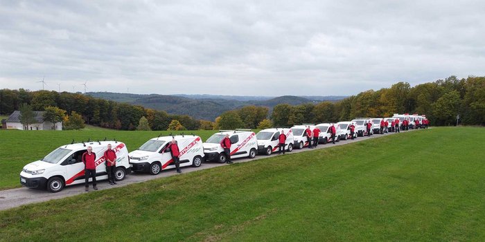 Eine Reihe von Lieferwagen steht auf einer Wiese, während Mitarbeiter in roten Jacken auf dem Weg posieren.