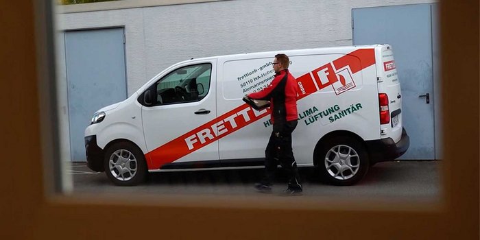 Ein Handwerker in schwarzer Arbeitskleidung trägt ein Werkzeug in der Hand, während er neben einem Firmenfahrzeug steht.