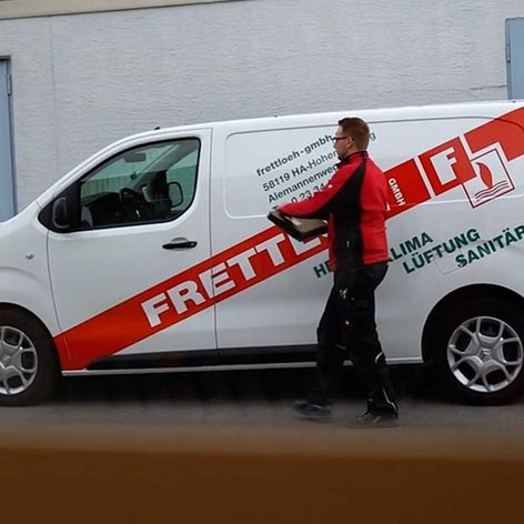 Ein Handwerker in schwarzer Arbeitskleidung trägt ein Werkzeug in der Hand, während er neben einem Firmenfahrzeug steht.