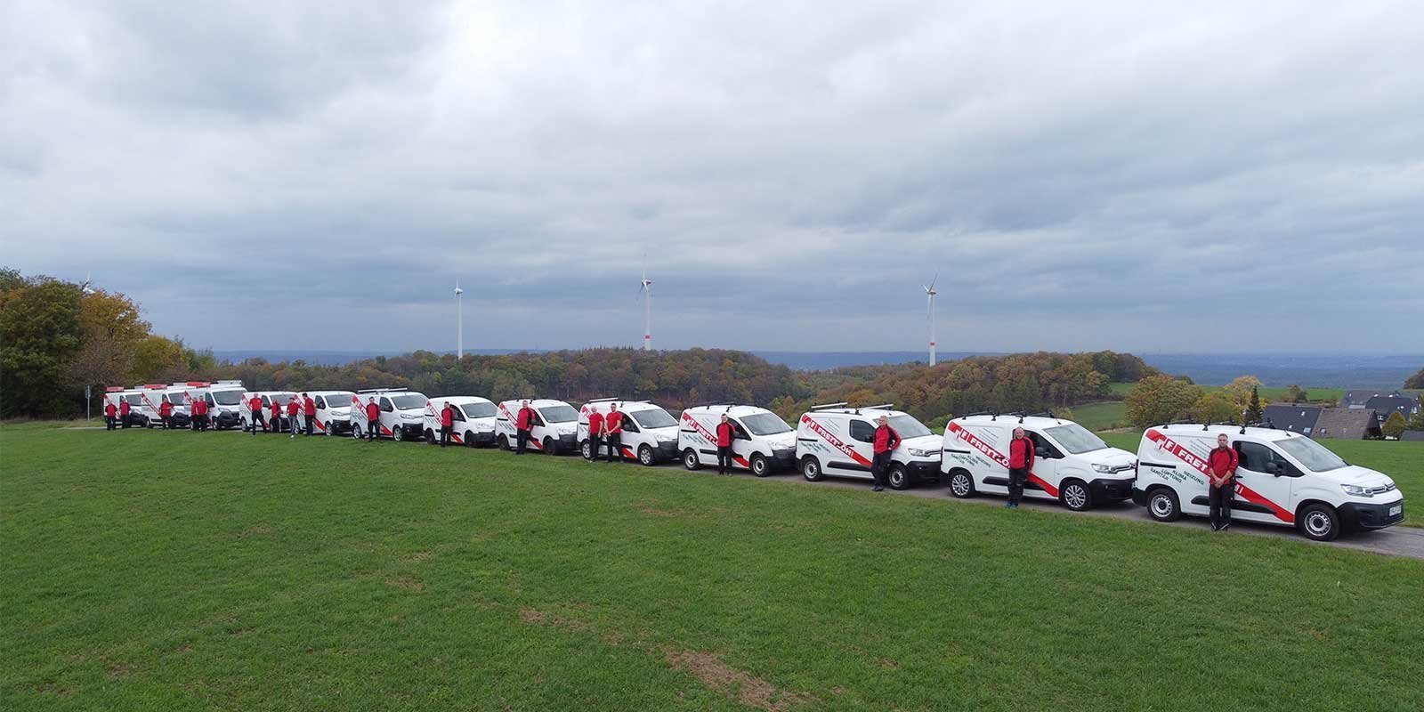 Eine Gruppe von Mitarbeitern in roten Jacken steht neben einer Reihe von weißen Lieferwagen auf einer grünen Wiese.