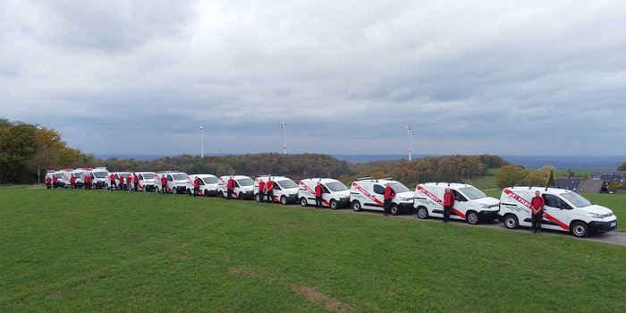 Eine Gruppe von Mitarbeitern in roten Jacken steht neben einer Reihe von weißen Lieferwagen auf einer grünen Wiese.