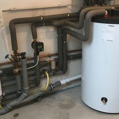 Ein Heizungsraum mit einem großen weißen Warmwasserspeicher und zahlreichen Rohren, die an die Wand angeschlossen sind.