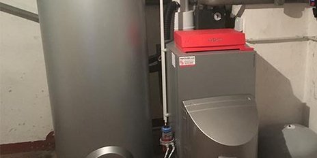 Ein silberner Warmwasserbereiter steht neben einem Heizkessel in einem Keller mit grauen Wänden und einem roten Boden.