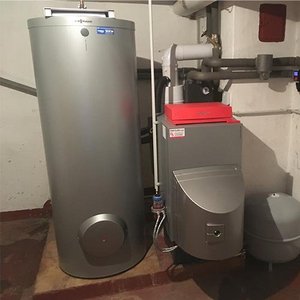 Ein silberner Warmwasserbereiter steht neben einem Heizkessel in einem Keller mit grauen Wänden und einem roten Boden.