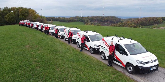 Eine Reihe von weißen Lieferwagen steht auf einer grünen Wiese, während Mitarbeiter in roten Shirts daneben posieren.