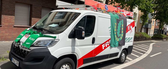 Ein weißer Lieferwagen mit bunten Fahnen und grünen Schals, die auf der Motorhaube drapiert sind, steht an einer Straße.