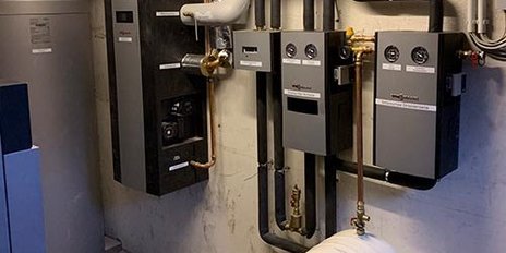 Die Wand ist mit verschiedenen Heizungsanlagen und Rohren ausgestattet, während ein großer, runder Wassertank im Raum steht.