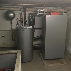 Ein Heizungsraum mit einem großen, grauen Heizkessel und einem zylindrischen Warmwasserspeicher, umgeben von Rohren.
