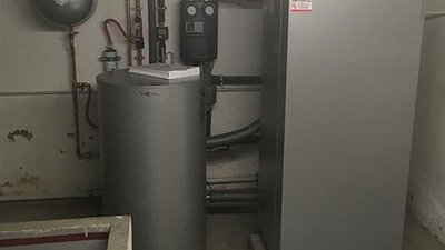 Ein Heizungsraum mit einem großen, grauen Heizkessel und einem zylindrischen Warmwasserspeicher, umgeben von Rohren.